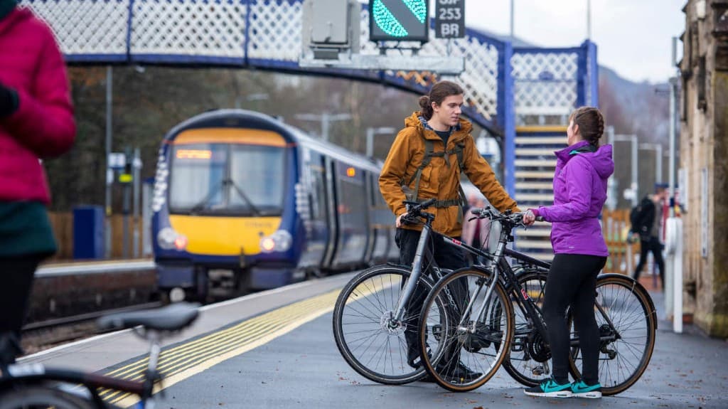 Two cyclists with bikes on a station platform as a train arrives, illustrating combined cycling and rail travel